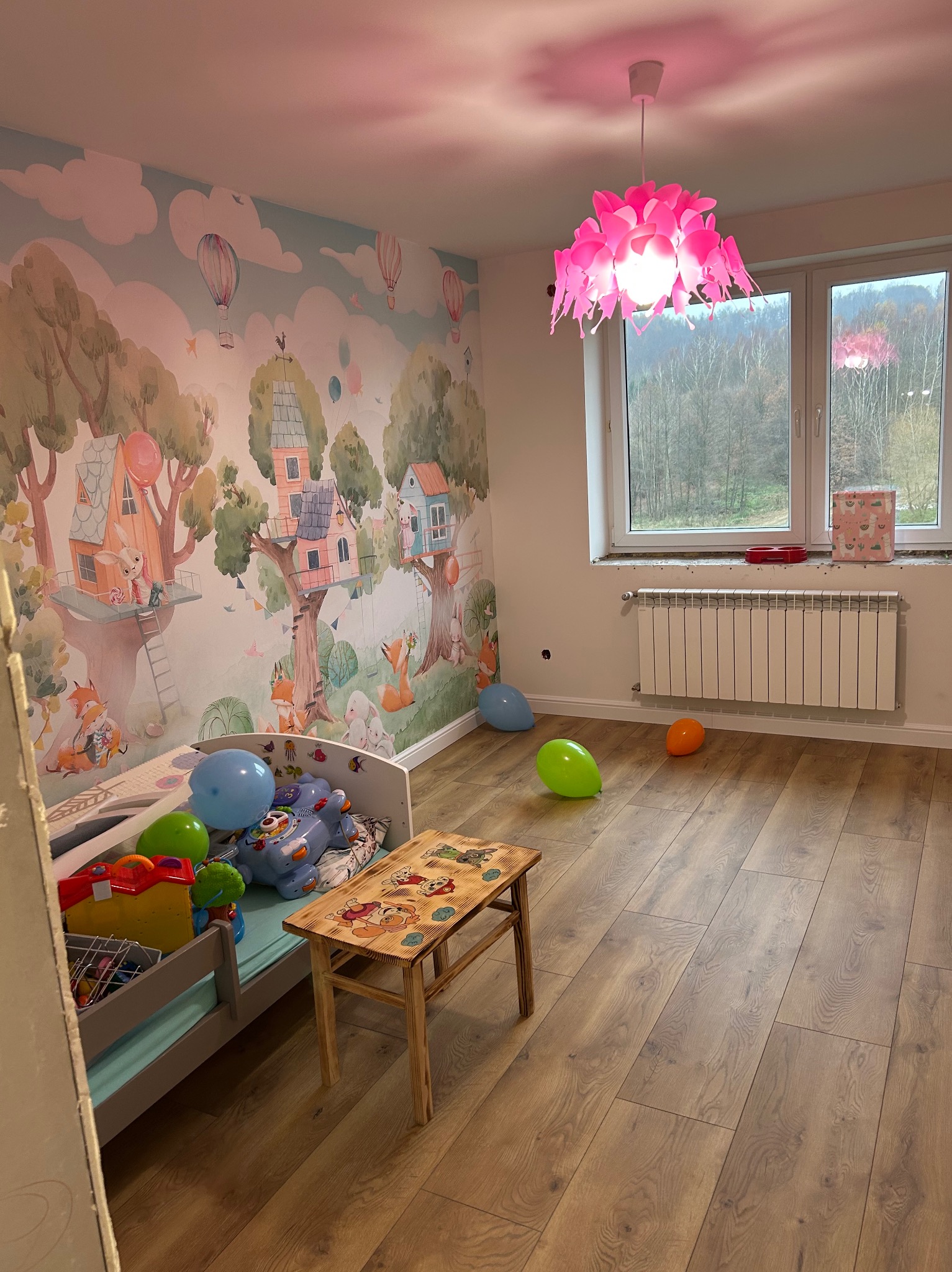 Pokój dziecięcy z tapetą w leśny motyw, drewnianą podłogą, różowym żyrandolem i grzejnikiem pod oknem. Na podłodze leżą kolorowe balony.