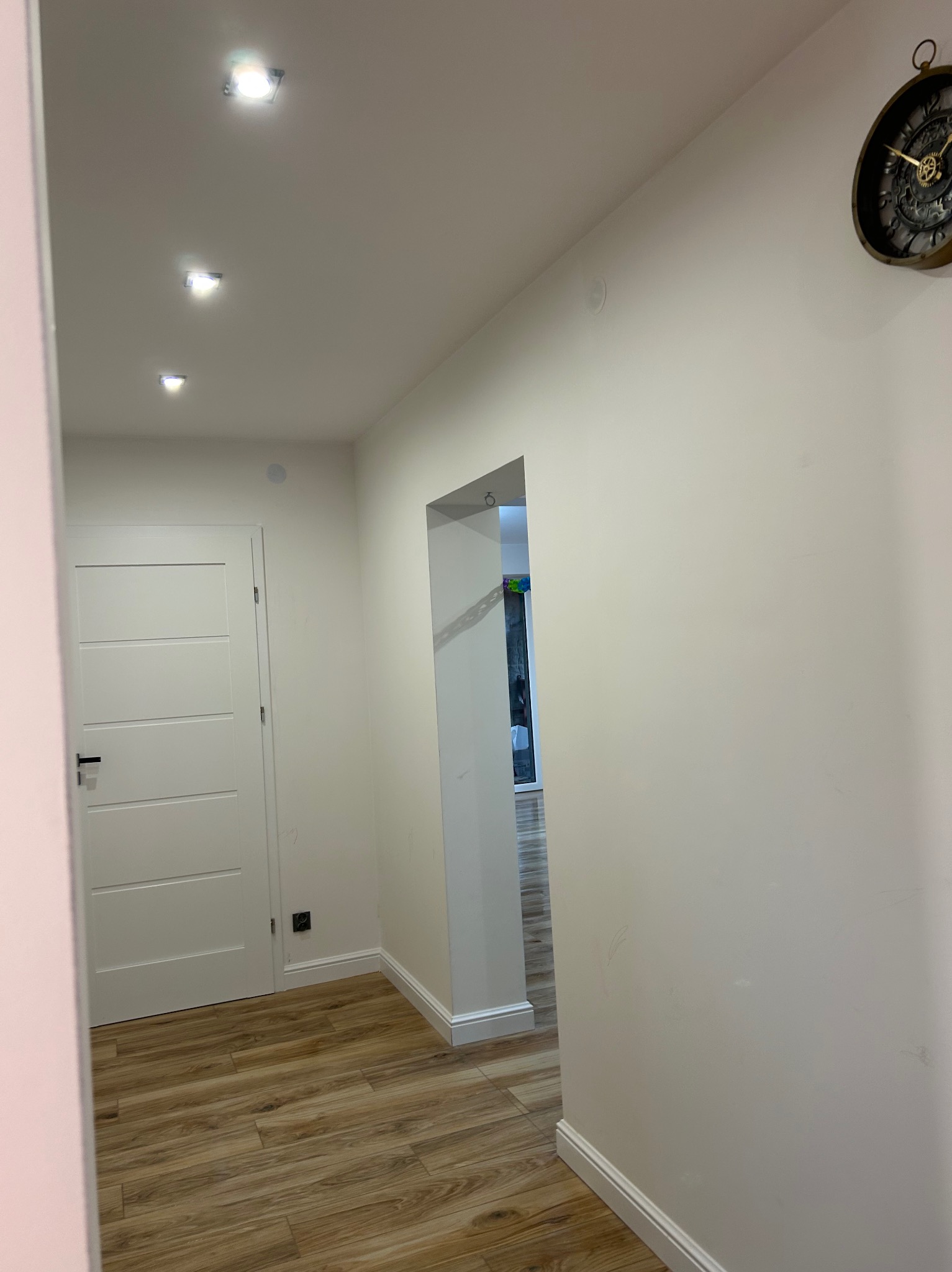 Korytarz po remoncie z panelami podłogowymi w odcieniach brązu, świeżo pomalowanymi na biało ścianami, białymi listwami przypodłogowymi, białymi drzwiami z czarną klamką i zegarem w stylu retro...