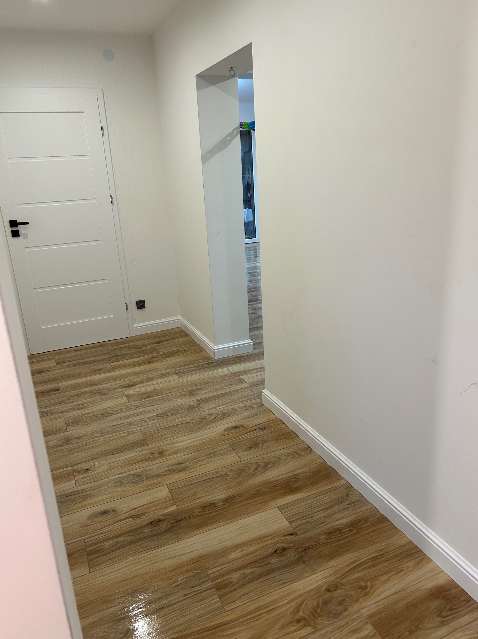 Korytarz z panelami podłogowymi imitującymi drewno, białe ściany z listwami przypodłogowymi, białe drzwi z czarną klamką i widok na dalszą część mieszkania.