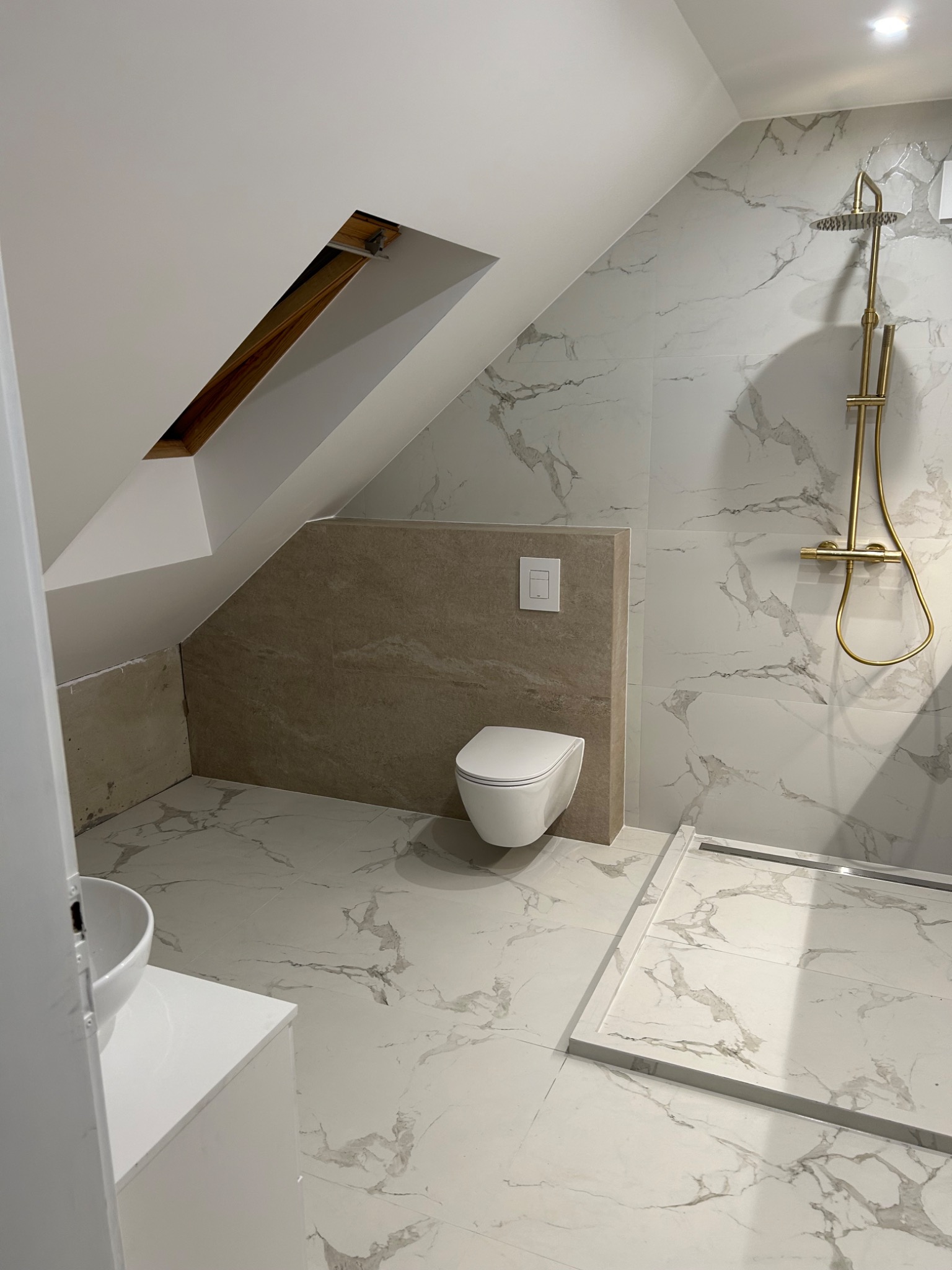 Wykończona łazienka na poddaszu z marmurowymi płytkami, złotą armaturą prysznicową i podwieszaną toaletą. Widoczny fragment umywalki i skos dachu.
