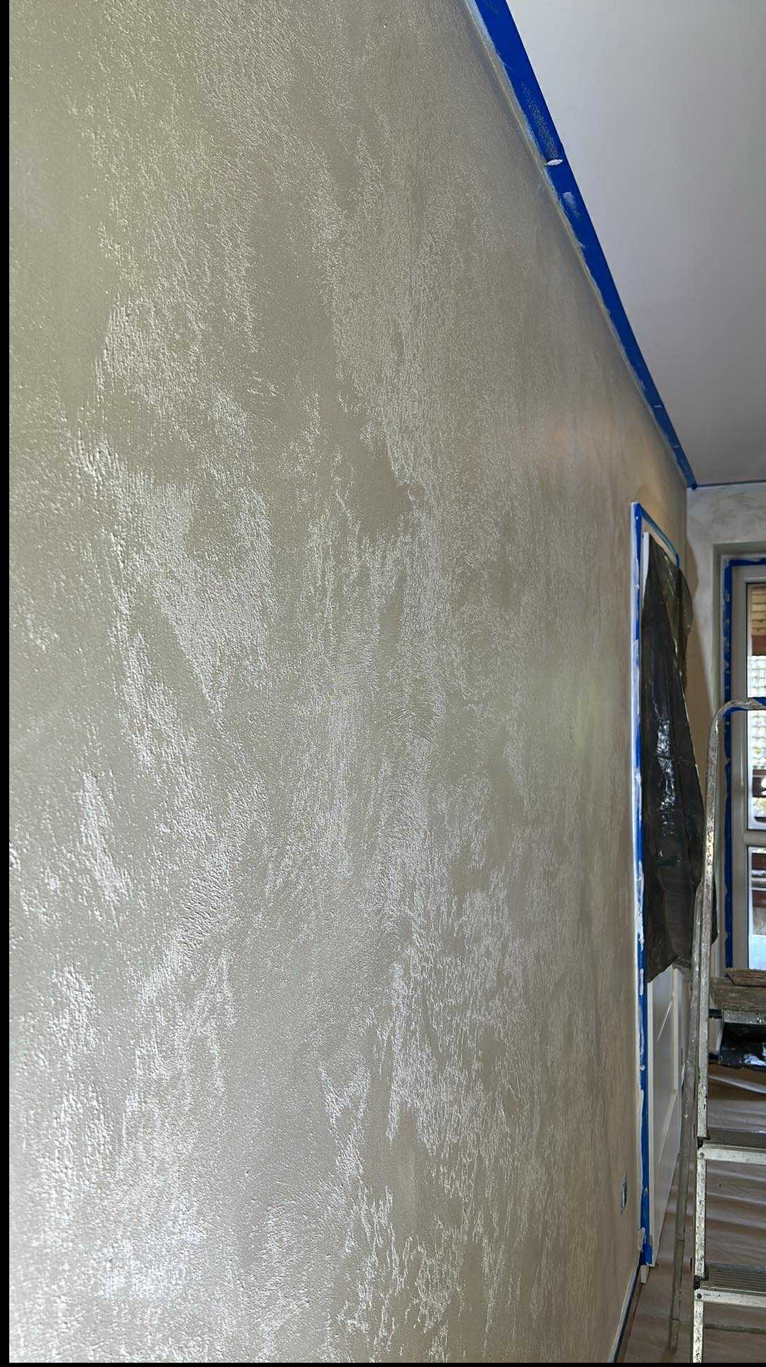 Ściana z dekoracyjną, srebrzystą fakturą, zabezpieczona niebieską taśmą malarską przy suficie i ościeżnicy. Widoczna drabina i okno w tle.