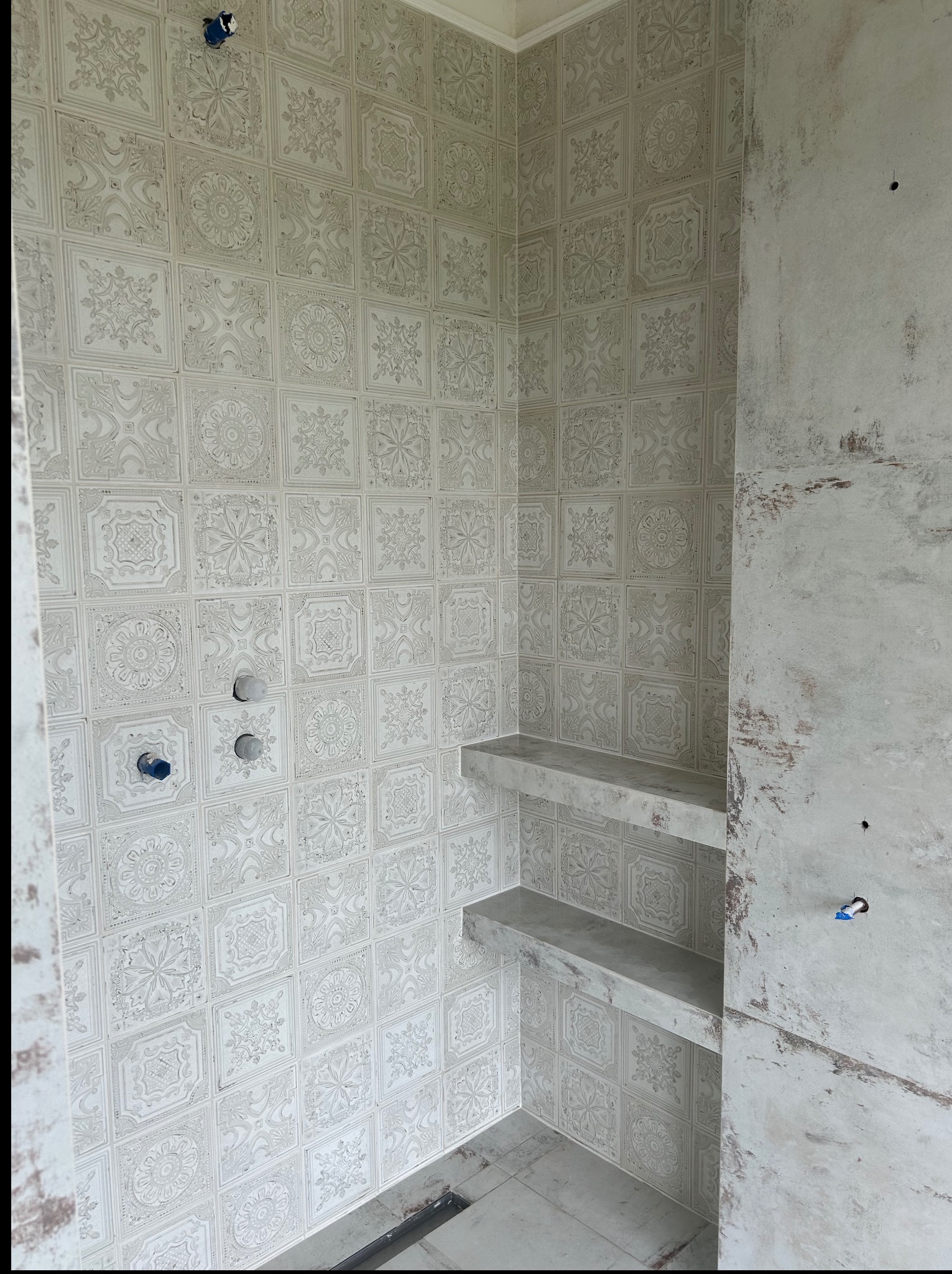 Wykończona ściana w łazience z dekoracyjnymi, jasnymi płytkami i wnękami. Surowa ściana obok, widoczne instalacje. Minimalistyczny design, neutralna kolorystyka.