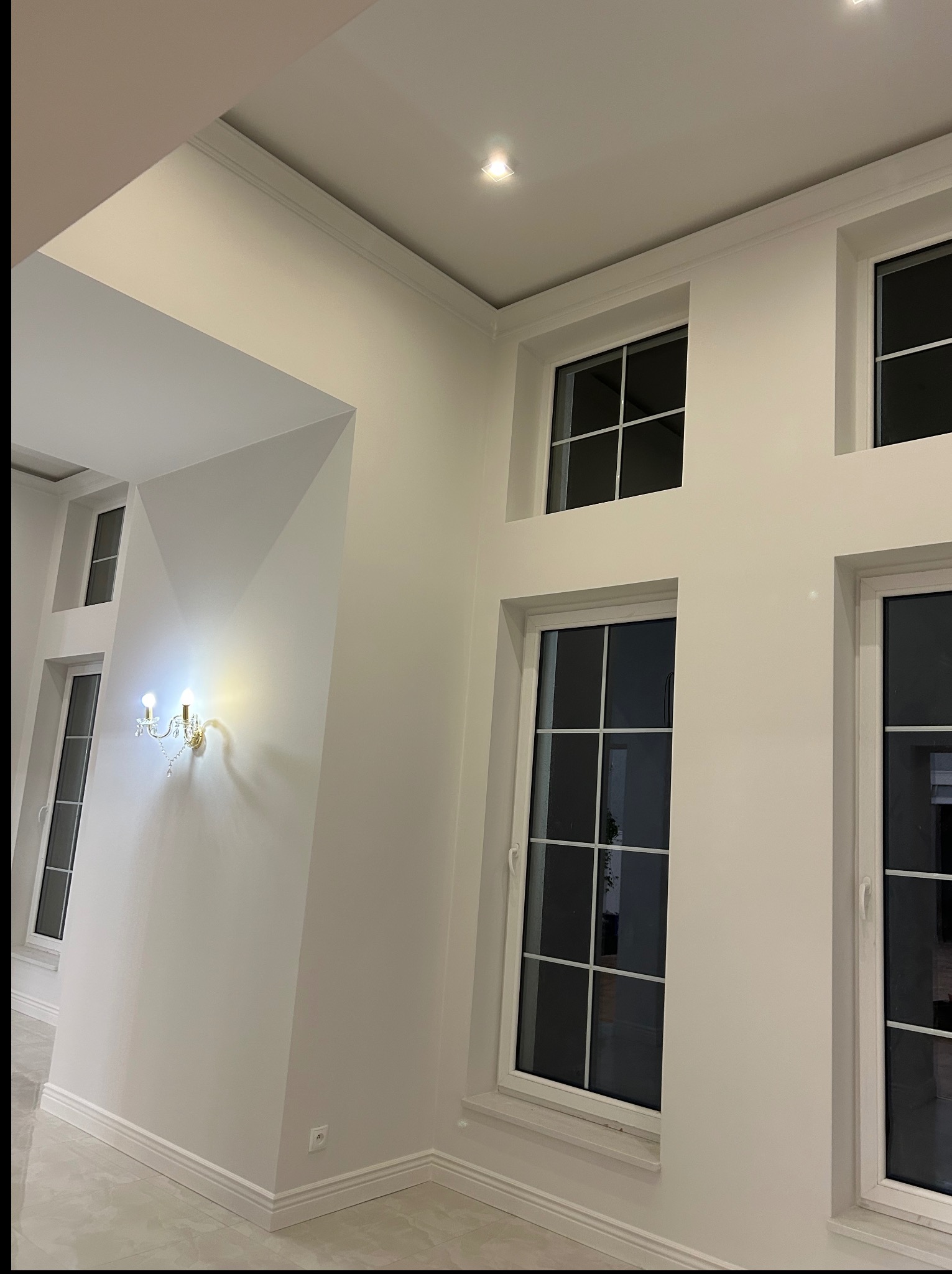 Jasne wnętrze po remoncie: białe ściany z dekoracyjną sztukaterią, okna z białymi ramami i elegancki kinkiet. Widok na odnowione pomieszczenie z perspektywy podłogi.