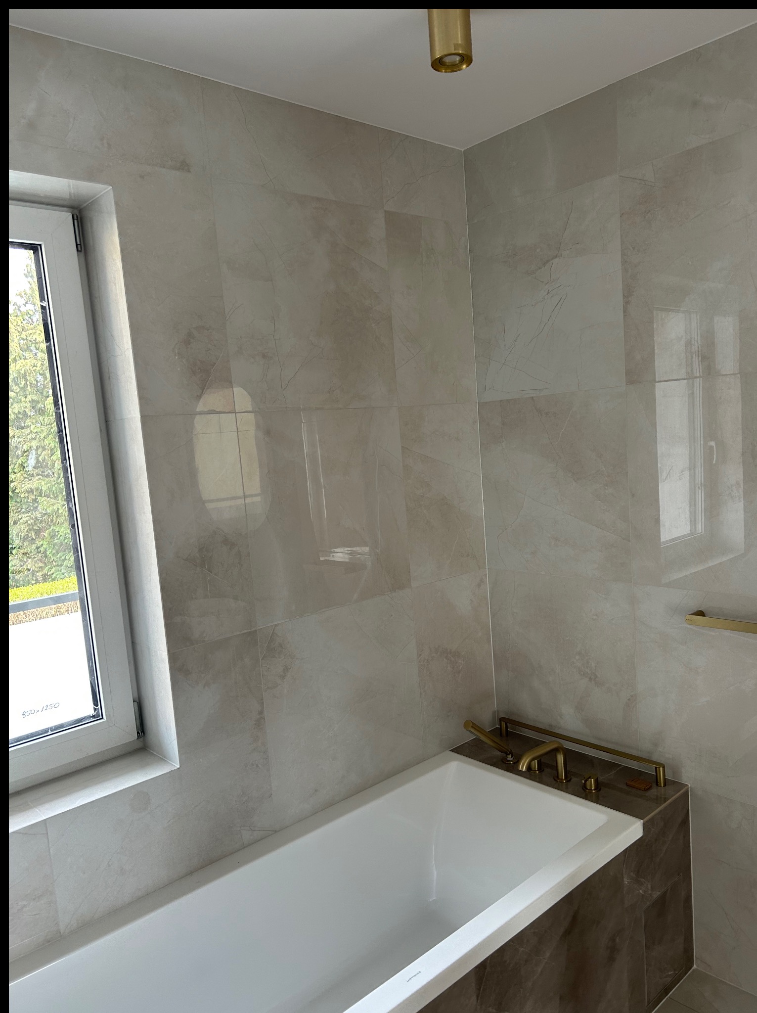 Nowoczesna łazienka po remoncie: biała wanna z brązowym obramowaniem, złota armatura, beżowe płytki imitujące marmur, okno z widokiem na zieleń. Minimalistyczny design.