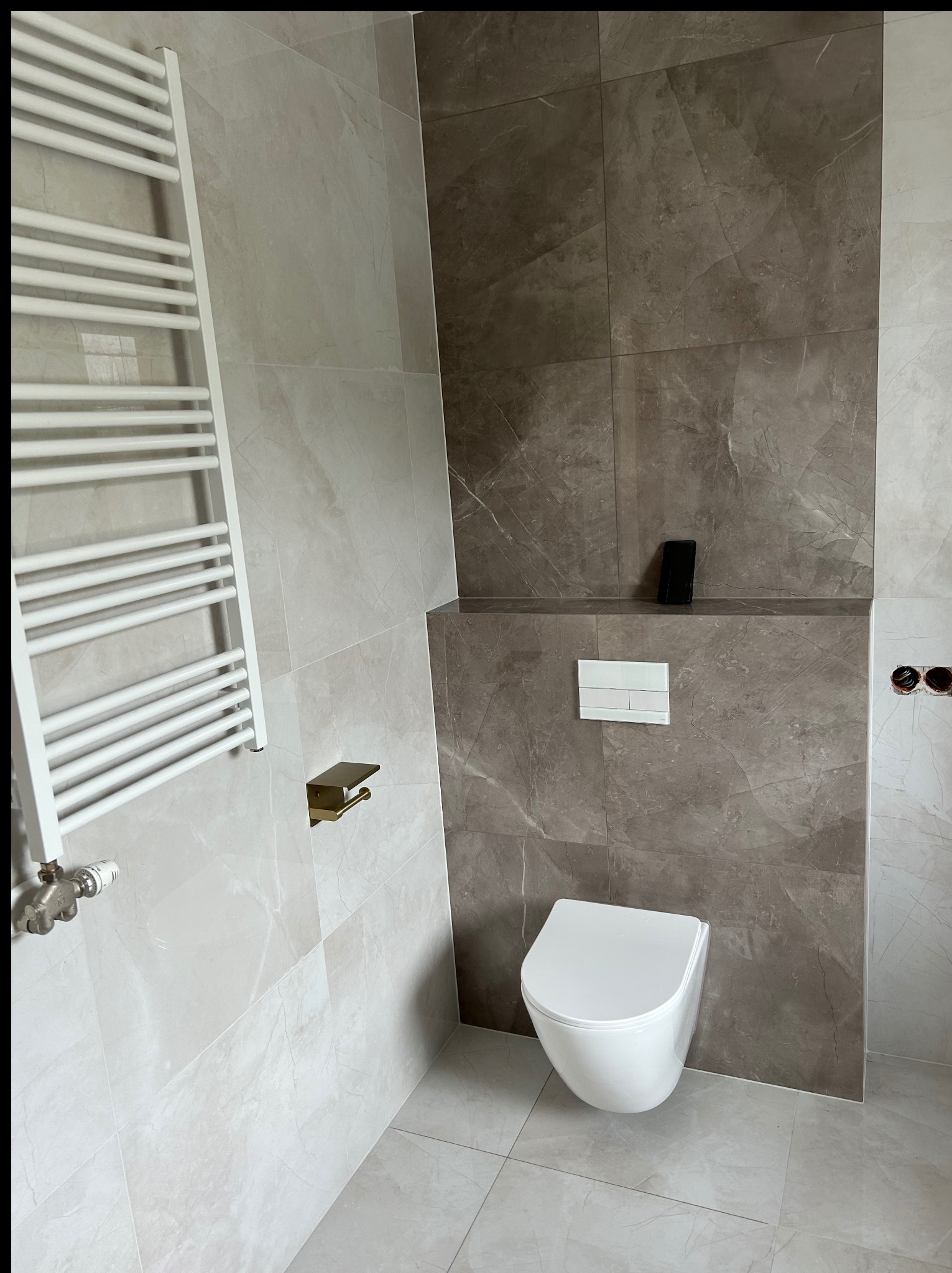 Nowoczesna łazienka z marmurowymi płytkami w odcieniach szarości i beżu, wisząca toaleta, grzejnik drabinkowy i złote akcenty. Minimalistyczny design.