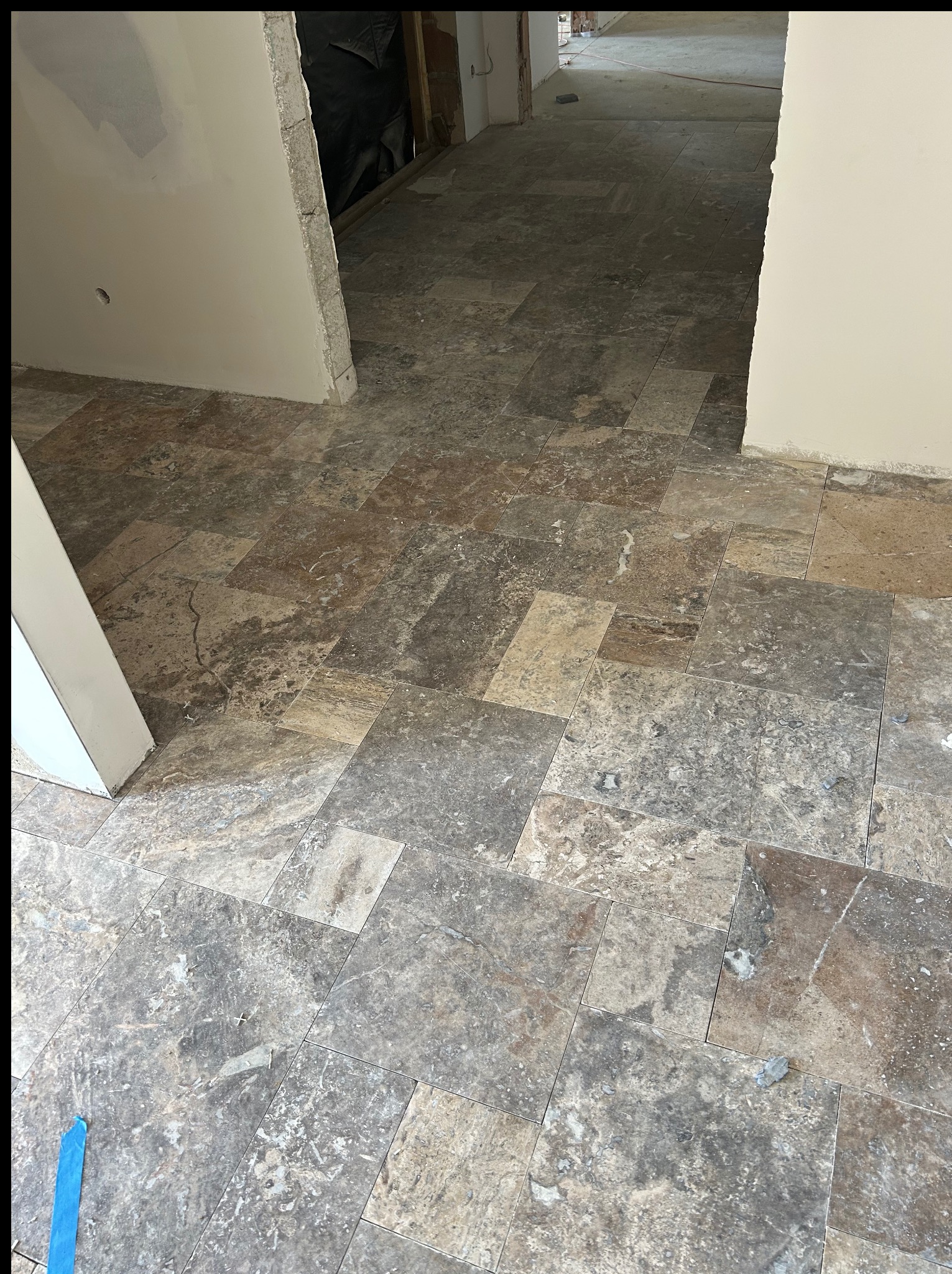 Ułożona podłoga z płytek o nieregularnym wzorze w odcieniach szarości i beżu, widok z góry, fragment ściany z widocznym otworem elektrycznym w tle.