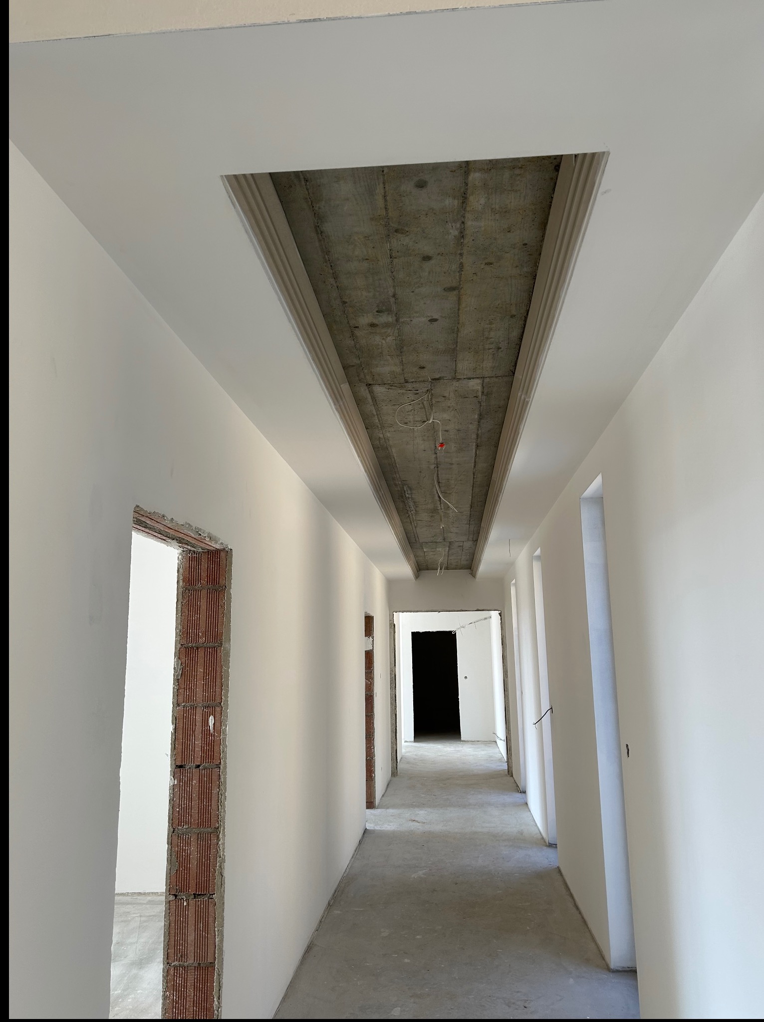 Korytarz w stanie deweloperskim z białymi ścianami i surowym betonowym stropem z dekoracyjną listwą. Widoczne otwory drzwiowe z ceglanym obramowaniem.