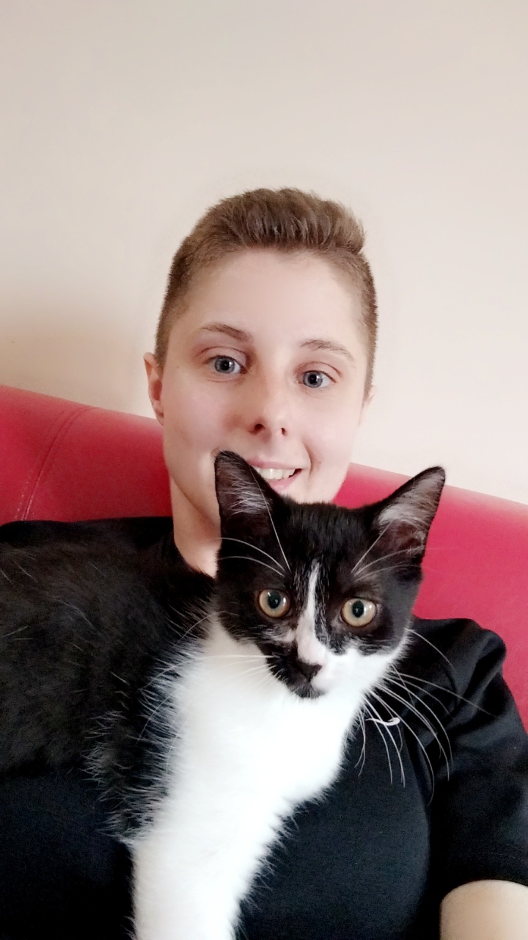 Portret uśmiechniętej kobiety z krótkimi włosami trzymającej czarno-białego kota na czerwonej kanapie.