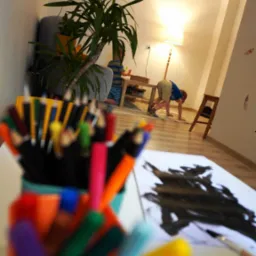 Dziecko maluje farbami na podłodze, obok stoi kubek z kredkami, w tle lampa podłogowa i roślina doniczkowa w salonie.