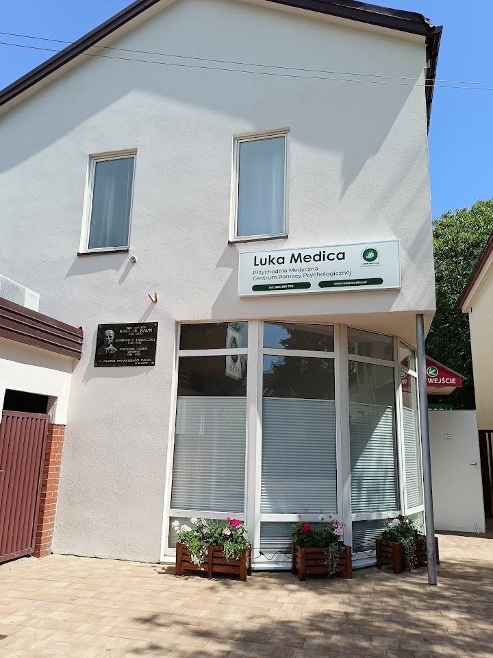 Fasada budynku z szyldem 'Luka Medica Przychodnia Medyczna Centrum Pomocy Psychologicznej', okna z żaluzjami i skrzynki z kwiatami.
