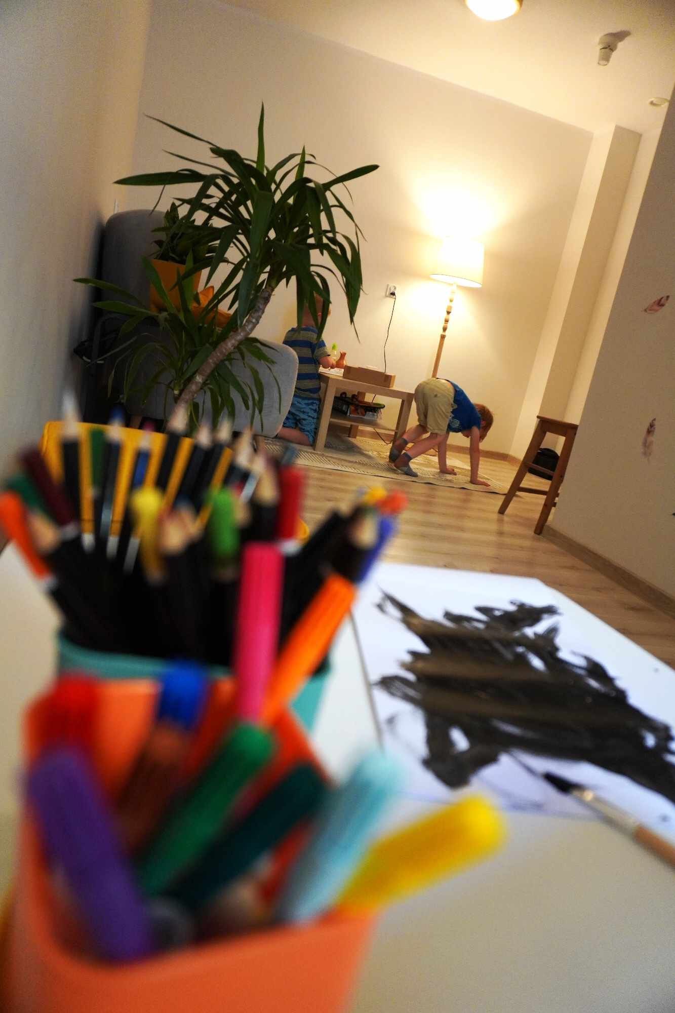 Dziecko maluje farbami na podłodze, obok stoi kubek z kredkami, w tle lampa podłogowa i roślina doniczkowa w salonie.