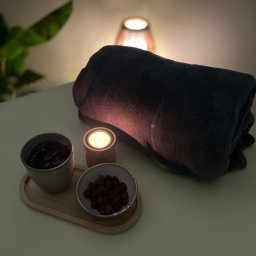 Krzysztof Masuje - Przytulne spa: zwinięty ciemny ręcznik, zapalona świeca w drewnianym świeczniku, miseczka z suszonymi owocami i kubek z naparem na drewnianej tacy, w tle ciepłe światło lampki.