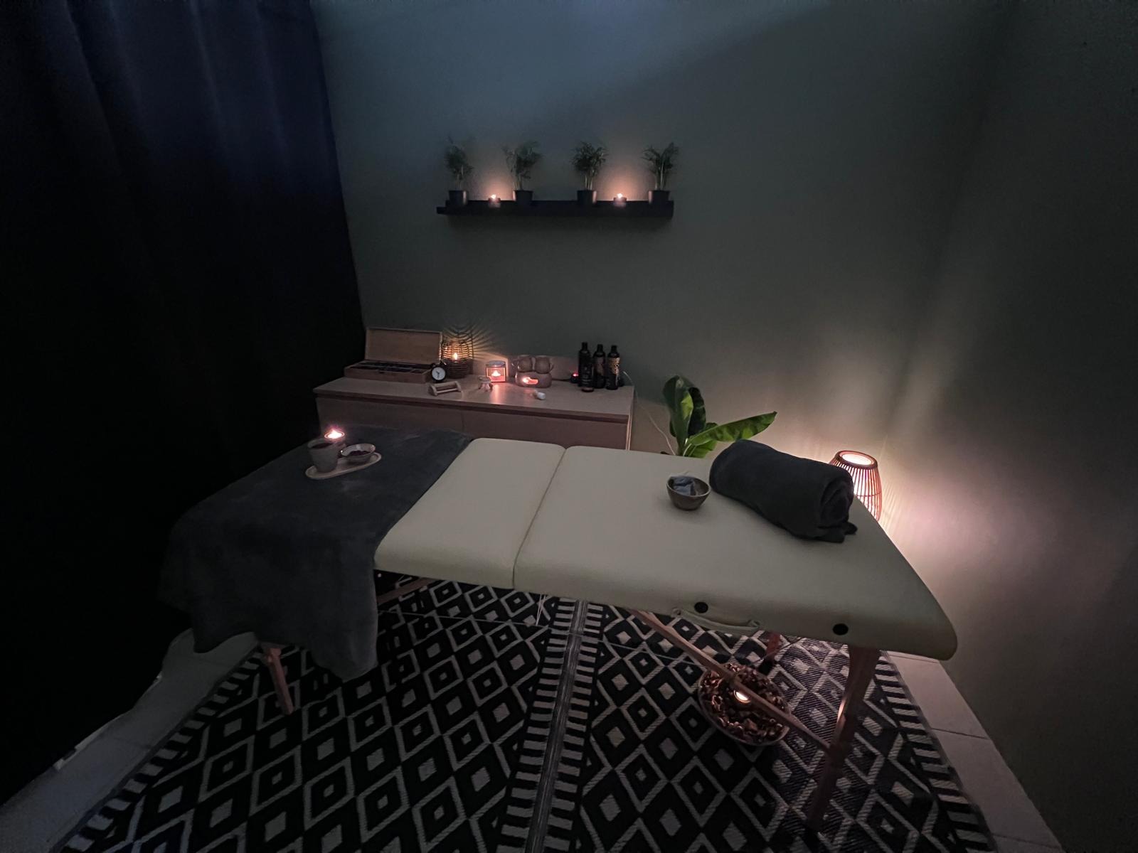 Przyciemnione wnętrze gabinetu z beżowym łóżkiem do zabiegów, czarno-białym dywanem w geometryczne wzory, świecami i roślinami w tle.