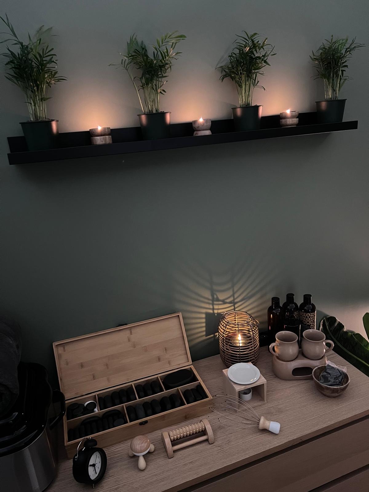 Przytulne wnętrze gabinetu: półka z roślinami i świecami, drewniana komoda z kamieniami do masażu w otwartym pudełku, podgrzewaczem, olejkami i akcesoriami do zabiegów.