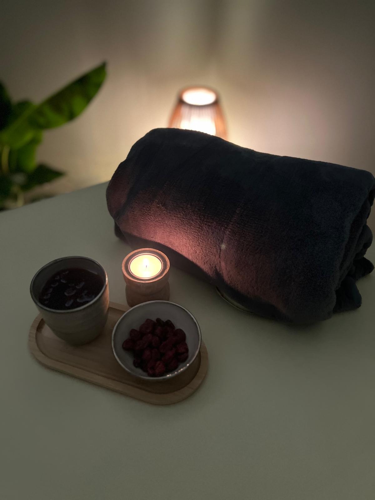 Przytulne spa: zwinięty ciemny ręcznik, zapalona świeca w drewnianym świeczniku, miseczka z suszonymi owocami i kubek z naparem na drewnianej tacy, w tle ciepłe światło lampki.
