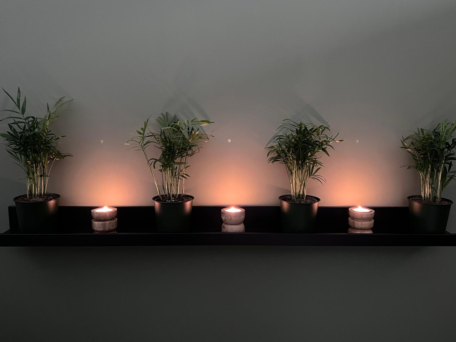Rząd doniczkowych palm arekowych i płonących świec w drewnianych podstawkach na czarnej półce przy szarej ścianie, tworzących relaksującą atmosferę.