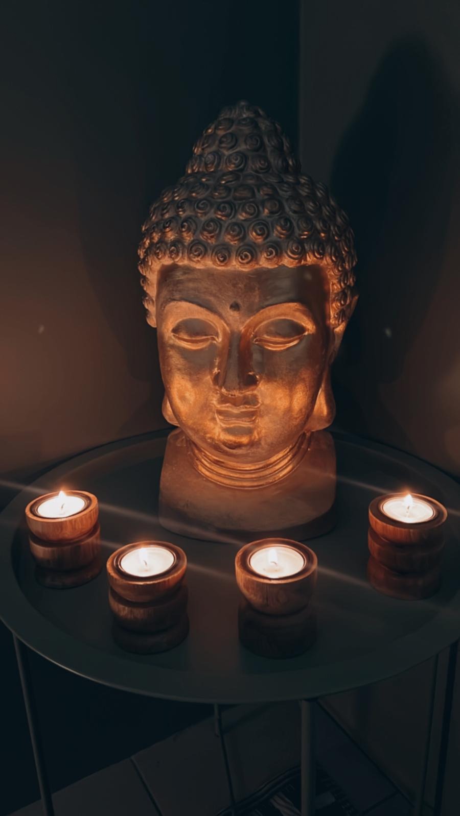 Głowa Buddy oświetlona świecami w drewnianych świecznikach na okrągłym stoliku, tworząca atmosferę relaksu i medytacji.
