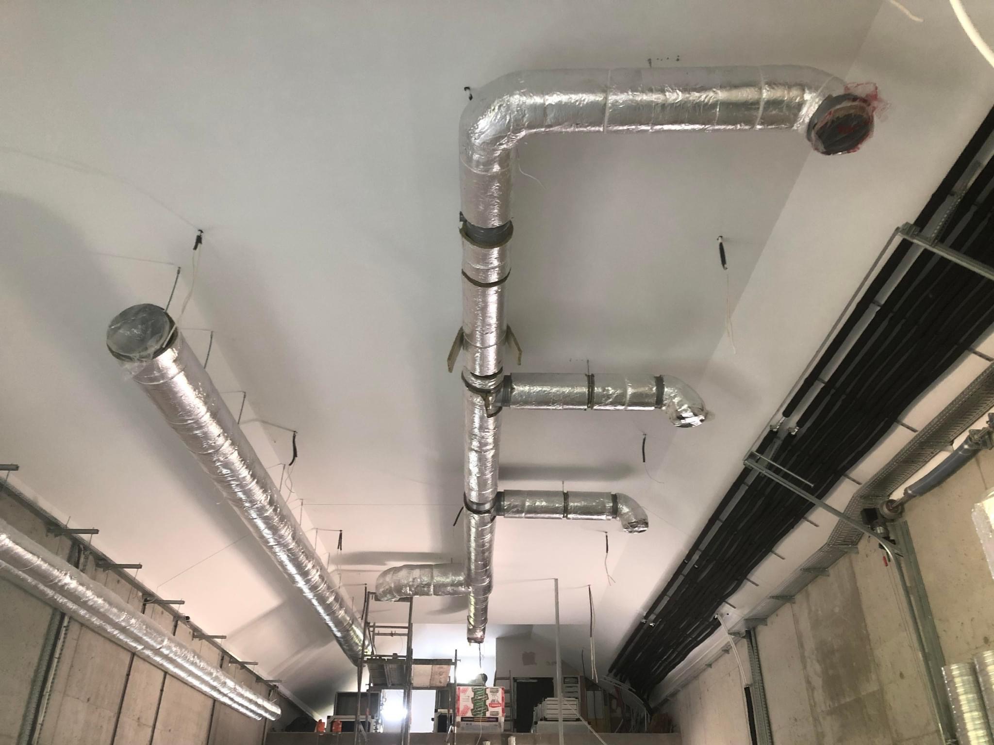 Instalacja kanałów wentylacyjnych z izolacją aluminiową, widok z dołu na sufit w surowym stanie budynku, z widocznymi przewodami elektrycznymi i elementami konstrukcyjnymi.