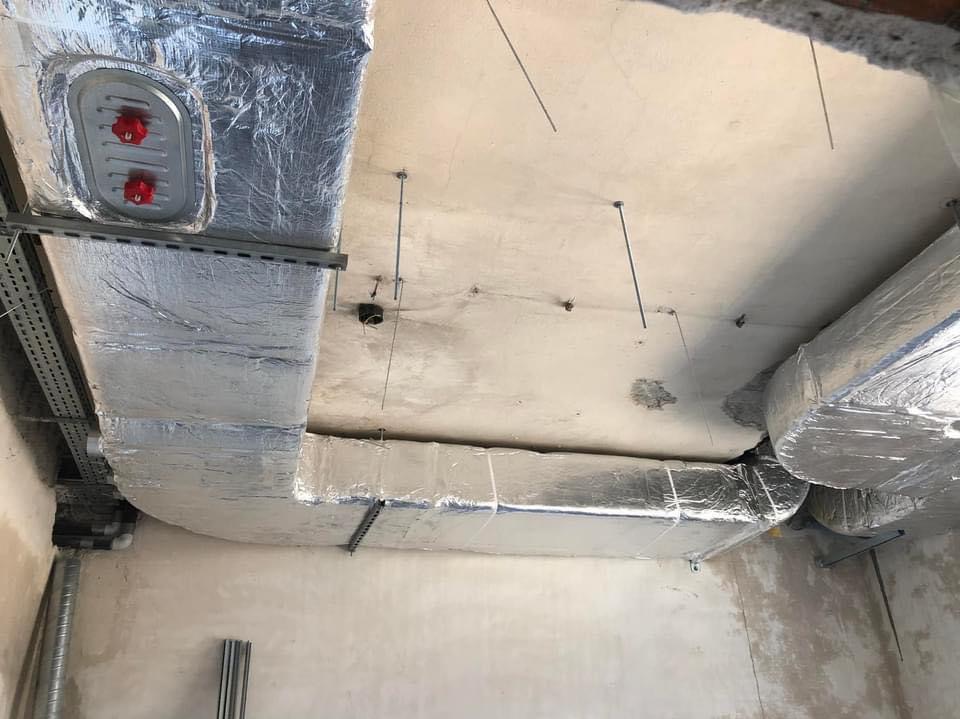 Instalacja kanałów wentylacyjnych z izolacją aluminiową na suficie budynku w stanie surowym, widoczne elementy montażowe i otwór w stropie.