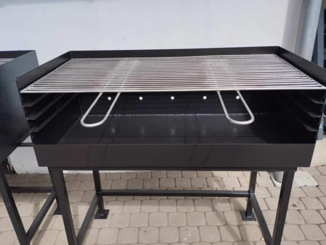 Czarny, metalowy grill ogrodowy z rusztem ze stali nierdzewnej, widoczne uchwyty do podnoszenia rusztu, ustawiony na kostce brukowej.