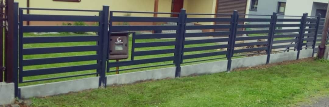 Nowoczesne ogrodzenie panelowe z poziomymi przęsłami w kolorze grafitowym, osadzone na betonowej podmurówce, z brązową skrzynką na listy z literą G, otoczone zielonym trawnikiem.