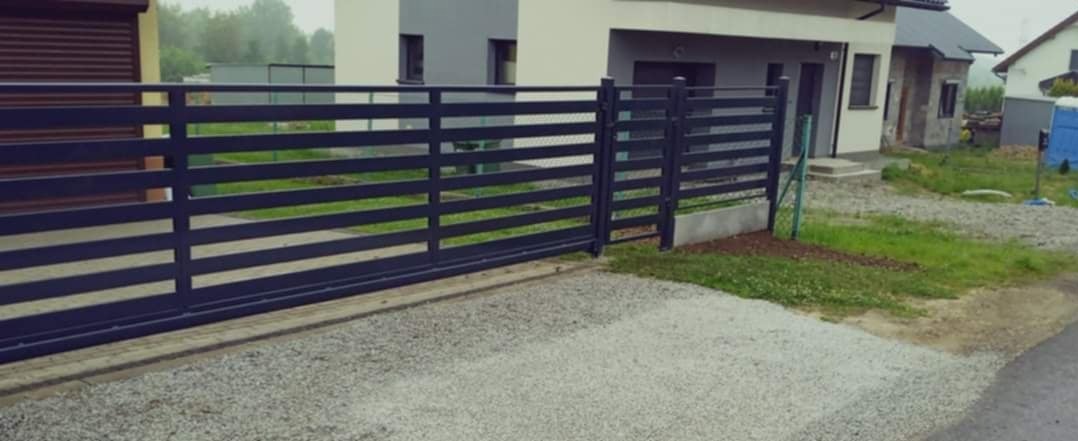 Nowoczesna, przesuwna brama wjazdowa z poziomych profili, pomalowana na ciemny kolor, na tle szarej nawierzchni i zabudowań.