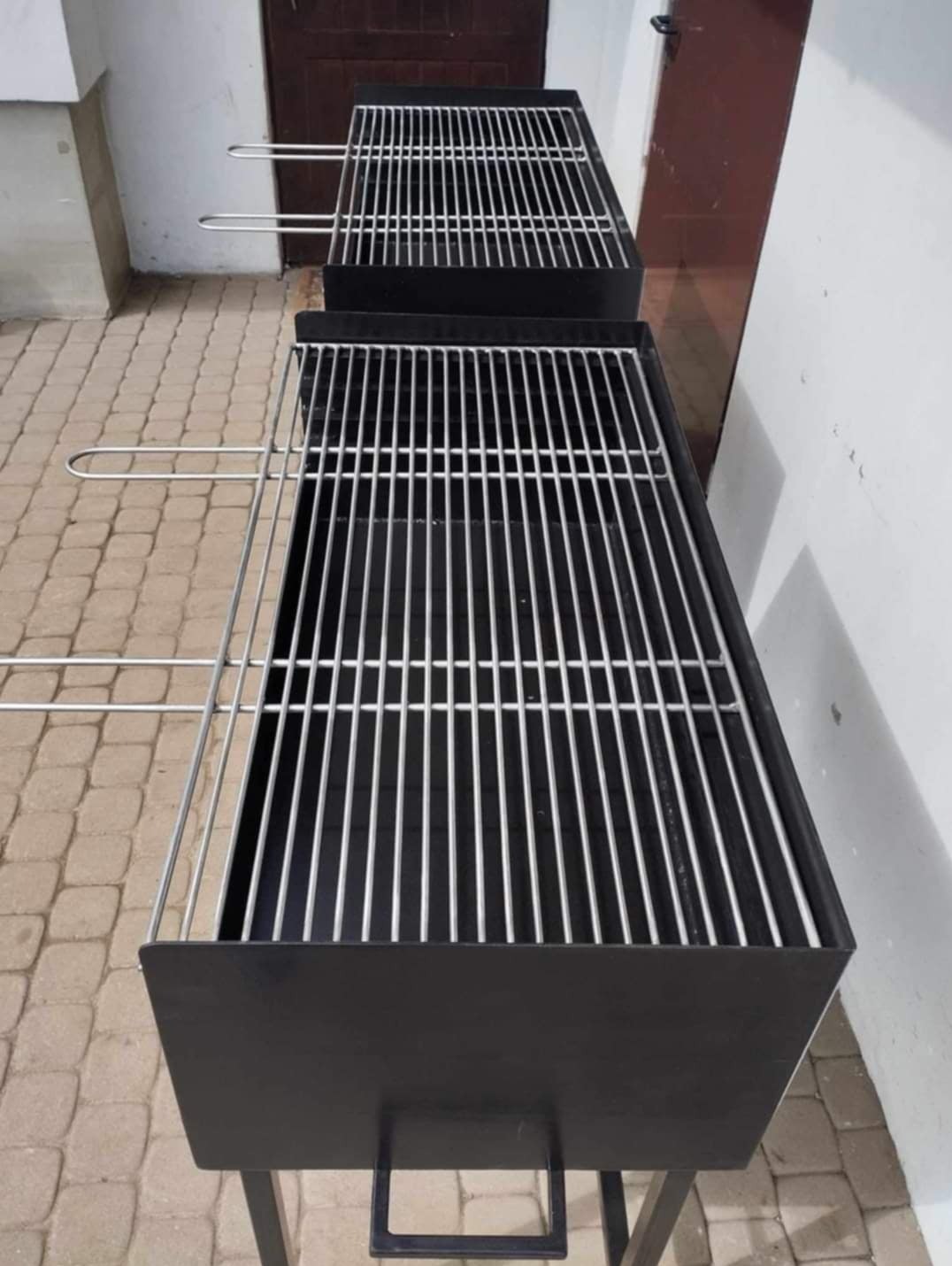 Dwa identyczne, czarne grille metalowe z rusztem ze stali nierdzewnej, ustawione jeden za drugim na kostce brukowej przy białej ścianie i brązowych drzwiach.