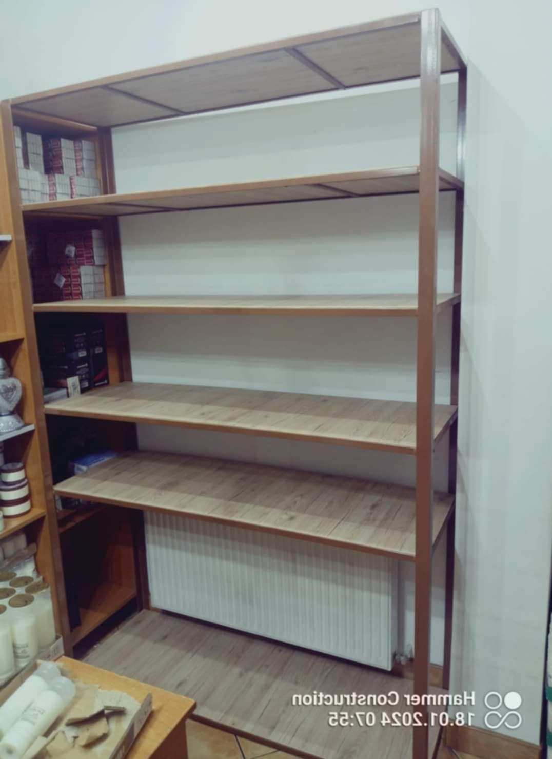 Metalowy regał z półkami imitującymi drewno, częściowo wypełniony przedmiotami, ustawiony na podłodze z panelami, z widocznym grzejnikiem w tle.