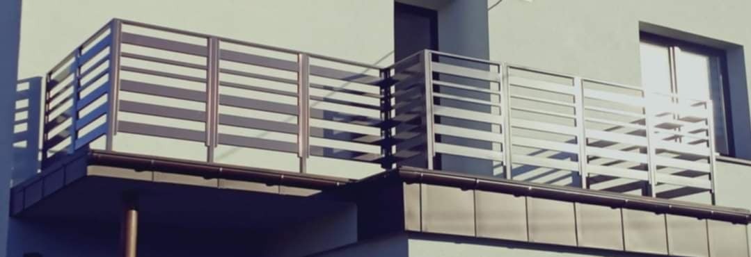 Nowoczesna balustrada balkonowa z poziomych, metalowych profili w kolorze szaro-srebrnym, zamontowana na budynku o jasnej elewacji.