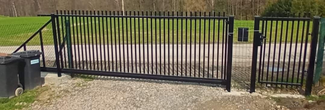 Czarna, przesuwna brama wjazdowa z pionowymi prętami, obok furtka z identycznym wzorem, na żwirowym podłożu, widoczne kosze na śmieci w tle.
