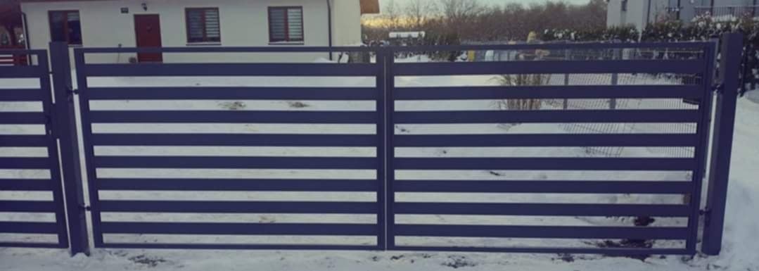 Nowoczesna, pozioma brama wjazdowa z metalowymi panelami w kolorze grafitowym na tle zasypanego śniegiem podwórka i dwukondygnacyjnego domu z jasną elewacją.