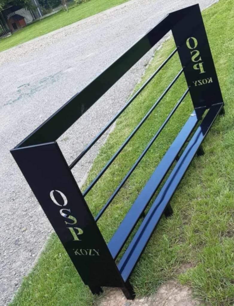 Czarny, metalowy stojak na siano z napisem 'OSP KOZY' w złotym kolorze, ustawiony na trawie obok asfaltowej drogi.