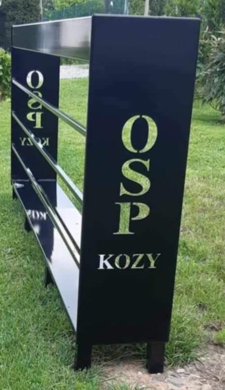 Czarny, metalowy regał na zewnątrz z napisem OSP KOZY, trzy poziomy z rurek.