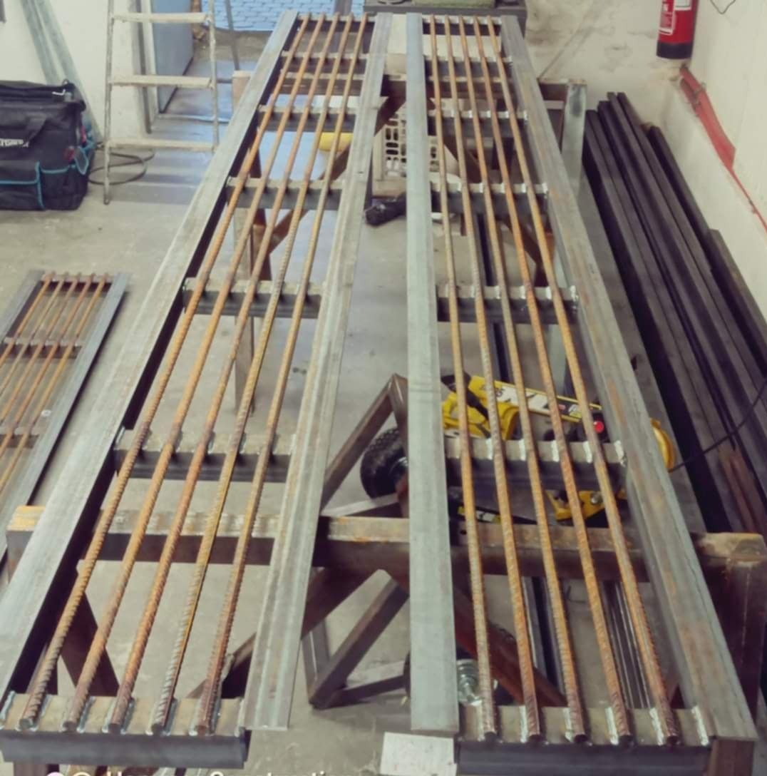 Dwa metalowe panele ogrodzeniowe z prętami, częściowo zmontowane na drewnianych kozłach, w warsztacie, z widoczną szlifierką kątową i zapasem profili.