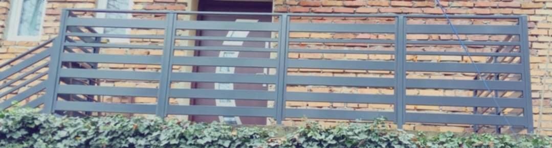 Szara, pozioma balustrada balkonowa z metalowych profili na tle ceglanej ściany budynku i zieleni bluszczu.