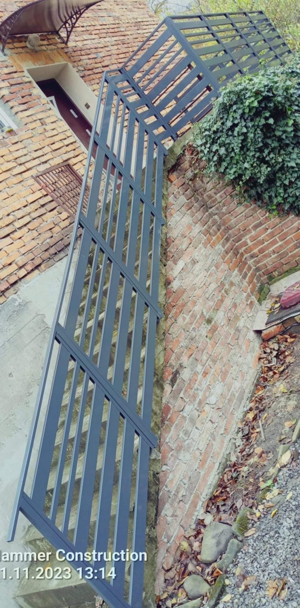 Szare, metalowe schody zewnętrzne z balustradą, prowadzące do domu z ceglanym murem i zielonym bluszczem.