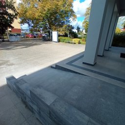 MA-KAM Remonty i Wykończenia Miłosz Kamiński - Wykończone wejście do budynku z nowym podjazdem dla niepełnosprawnych, widoczne szare płytki i struktura tynku na elewacji, kolumny budynku.