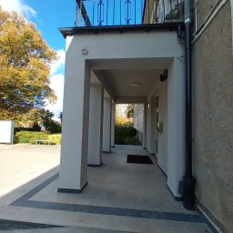 MA-KAM Remonty i Wykończenia Miłosz Kamiński - Odnowione wejście do budynku z kolumnadą, szara elewacja, widoczny fragment balkonu z czarną balustradą, na pierwszym planie nowa, szara płytka chodnikowa z czarnymi obramowaniami.
