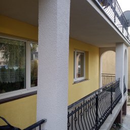 MA-KAM Remonty i Wykończenia Miłosz Kamiński - Żółta elewacja domu z białymi filarami i czarną, kutą balustradą balkonu, widok z boku, z fragmentem okna z firanką.