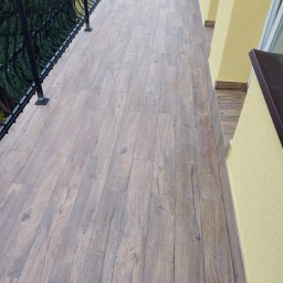 MA-KAM Remonty i Wykończenia Miłosz Kamiński - Balkon z podłogą wyłożoną panelami imitującymi drewno, z czarną, ozdobną balustradą i żółtą elewacją budynku.