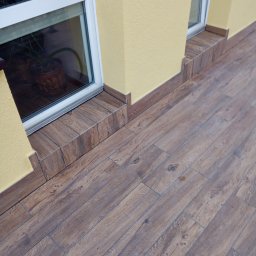 MA-KAM Remonty i Wykończenia Miłosz Kamiński - Elewacja budynku z oknem, wykończona płytkami imitującymi drewno na cokole i wokół okna. Żółty tynk strukturalny na ścianie. Widoczne łączenia płytek. Podłoga wyłożona panelami podłogowymi.