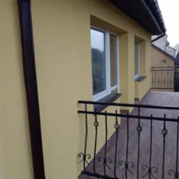 Żółta elewacja budynku z balkonem, brązowa rynna i balustrada balkonu z metalowymi zdobieniami.