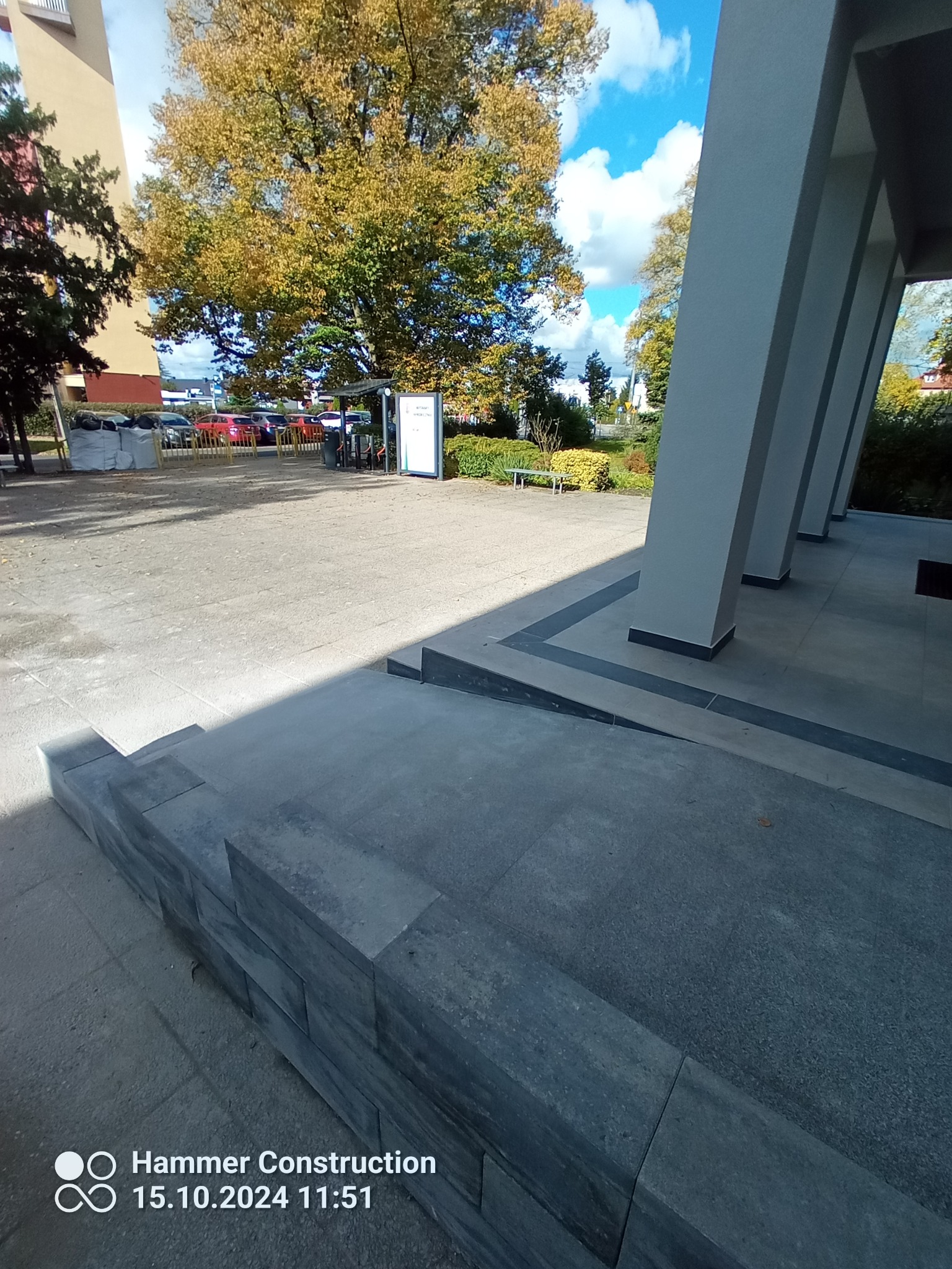 Wykończone wejście do budynku z nowym podjazdem dla niepełnosprawnych, widoczne szare płytki i struktura tynku na elewacji, kolumny budynku.