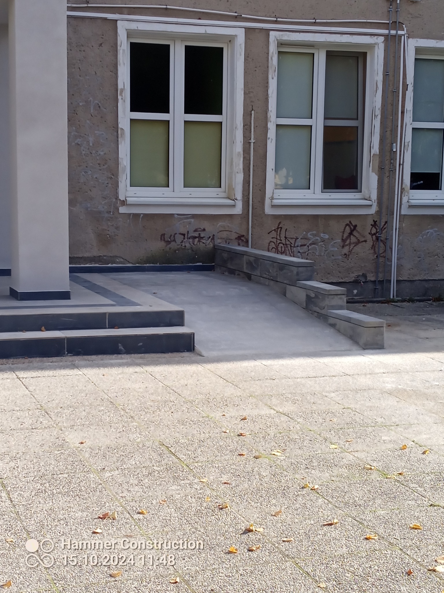 Rampa dla niepełnosprawnych z szarych płyt prowadząca do budynku z oknami i graffiti na ścianie, widok z zewnątrz.