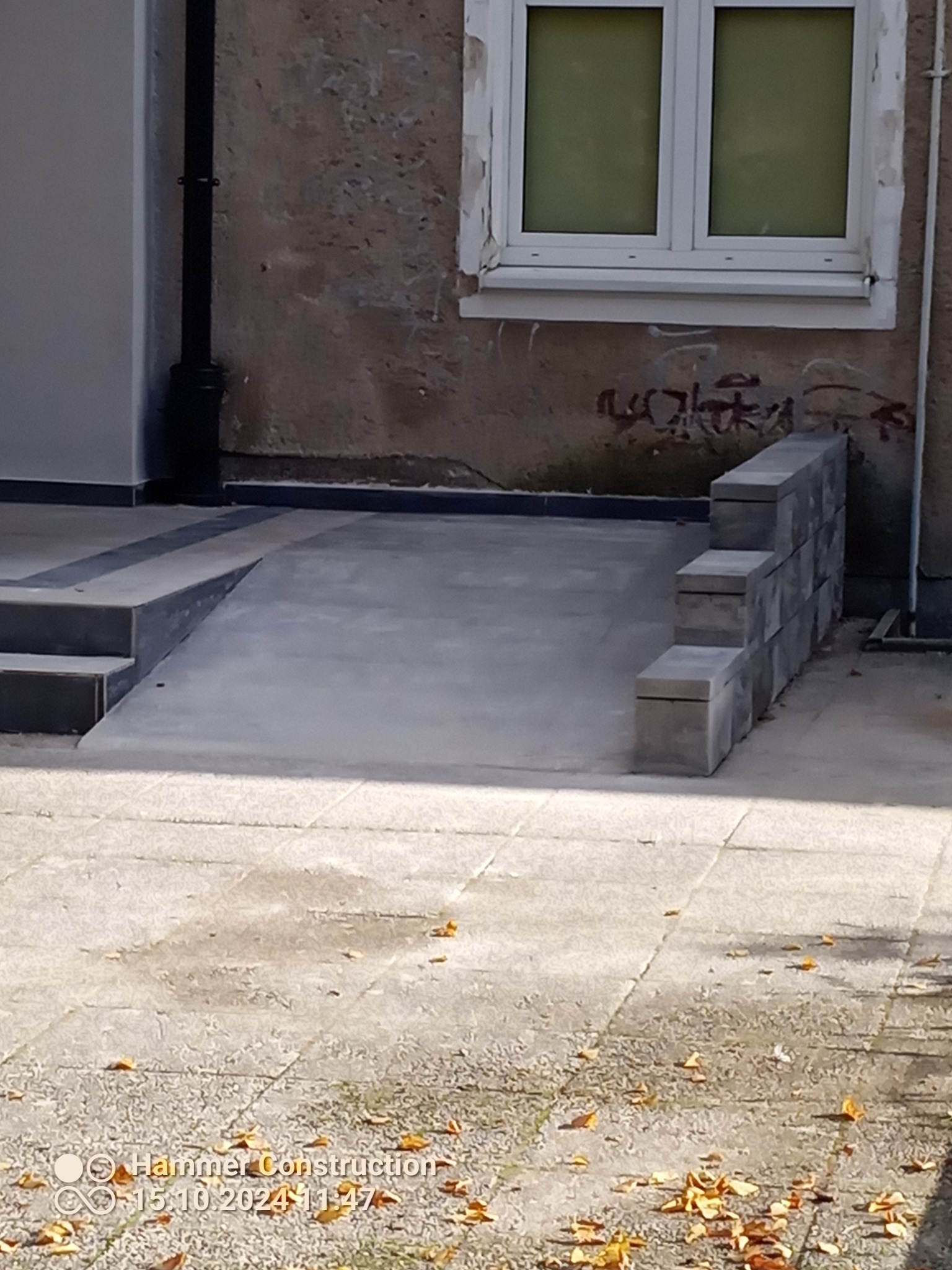Betonowy podjazd dla niepełnosprawnych z obudową z bloczków betonowych, wykonany przy budynku z widocznymi śladami graffiti i zniszczeń na elewacji.