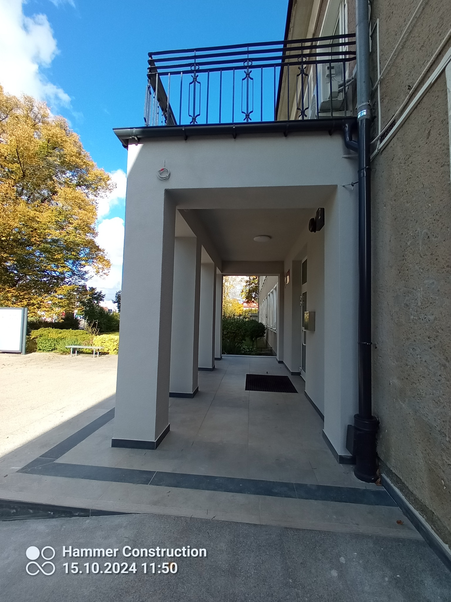Odnowione wejście do budynku z kolumnadą, szara elewacja, widoczny fragment balkonu z czarną balustradą, na pierwszym planie nowa, szara płytka chodnikowa z czarnymi obramowaniami.