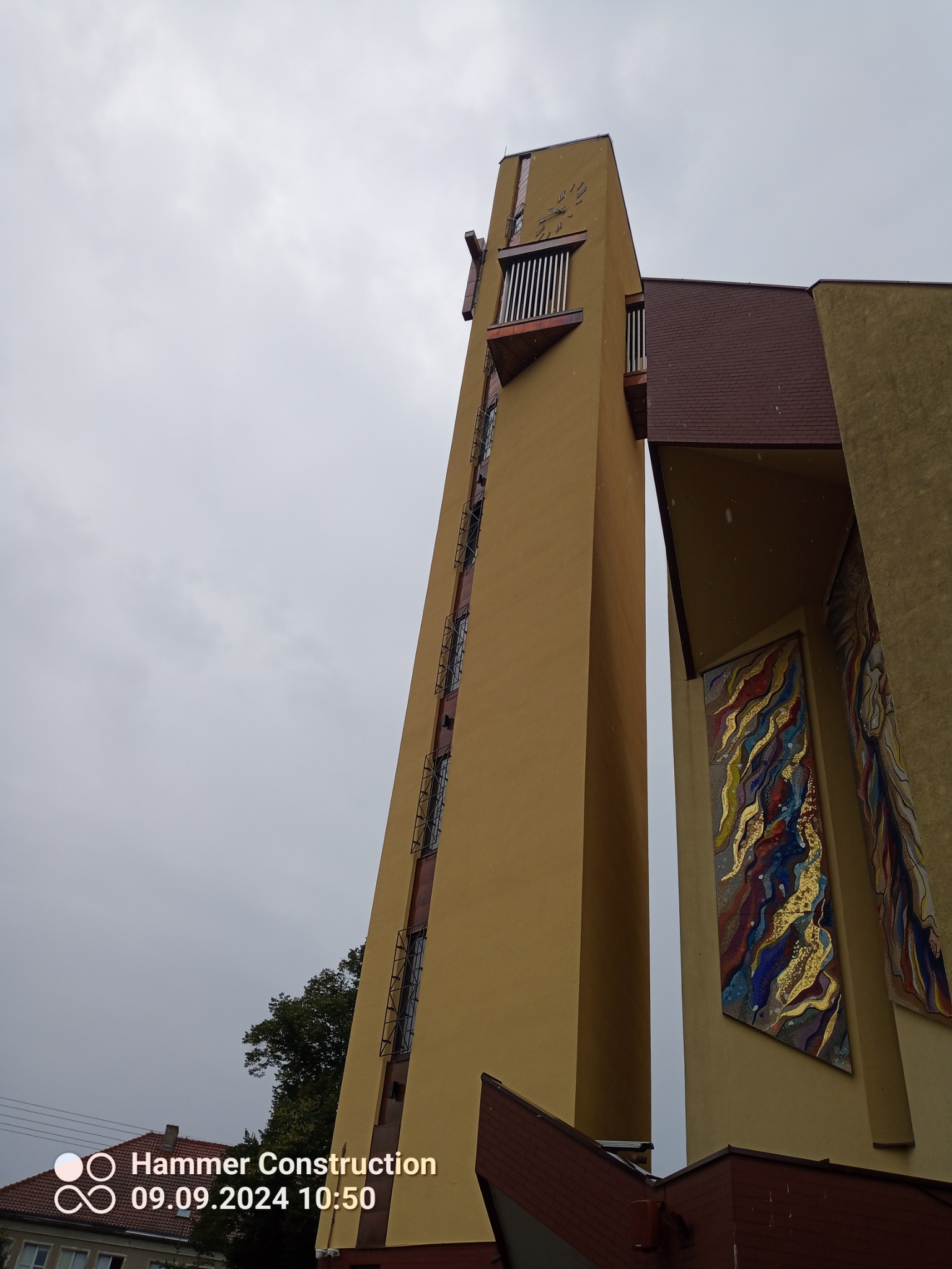 Wysoka, smukła wieża kościelna z zegarem i mozaikami na elewacji, sfotografowana z dołu w pochmurny dzień.