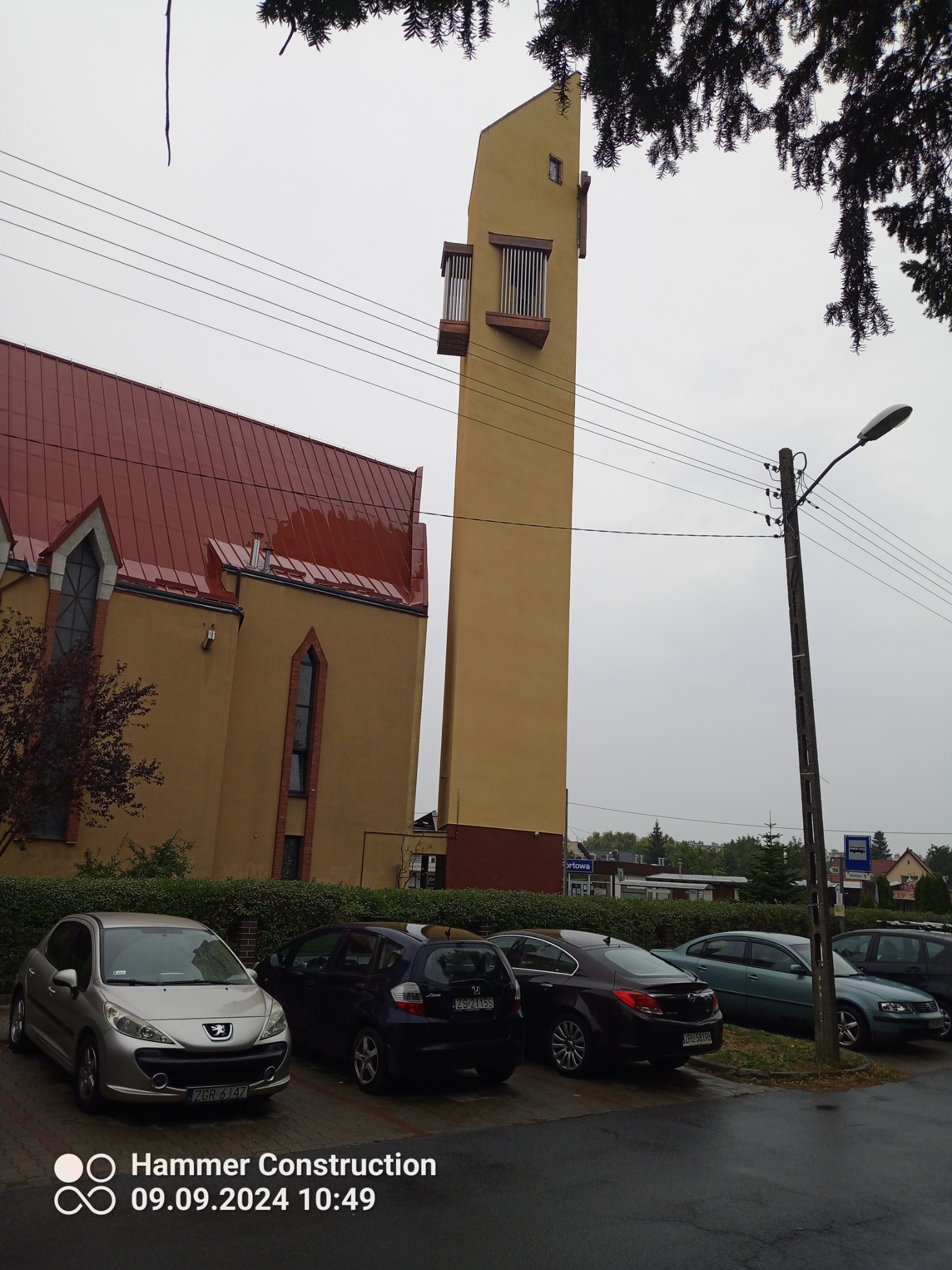 Żółty budynek kościoła z czerwoną dachówką i wysoką wieżą, widziany z poziomu ulicy z zaparkowanymi samochodami w pochmurny dzień.