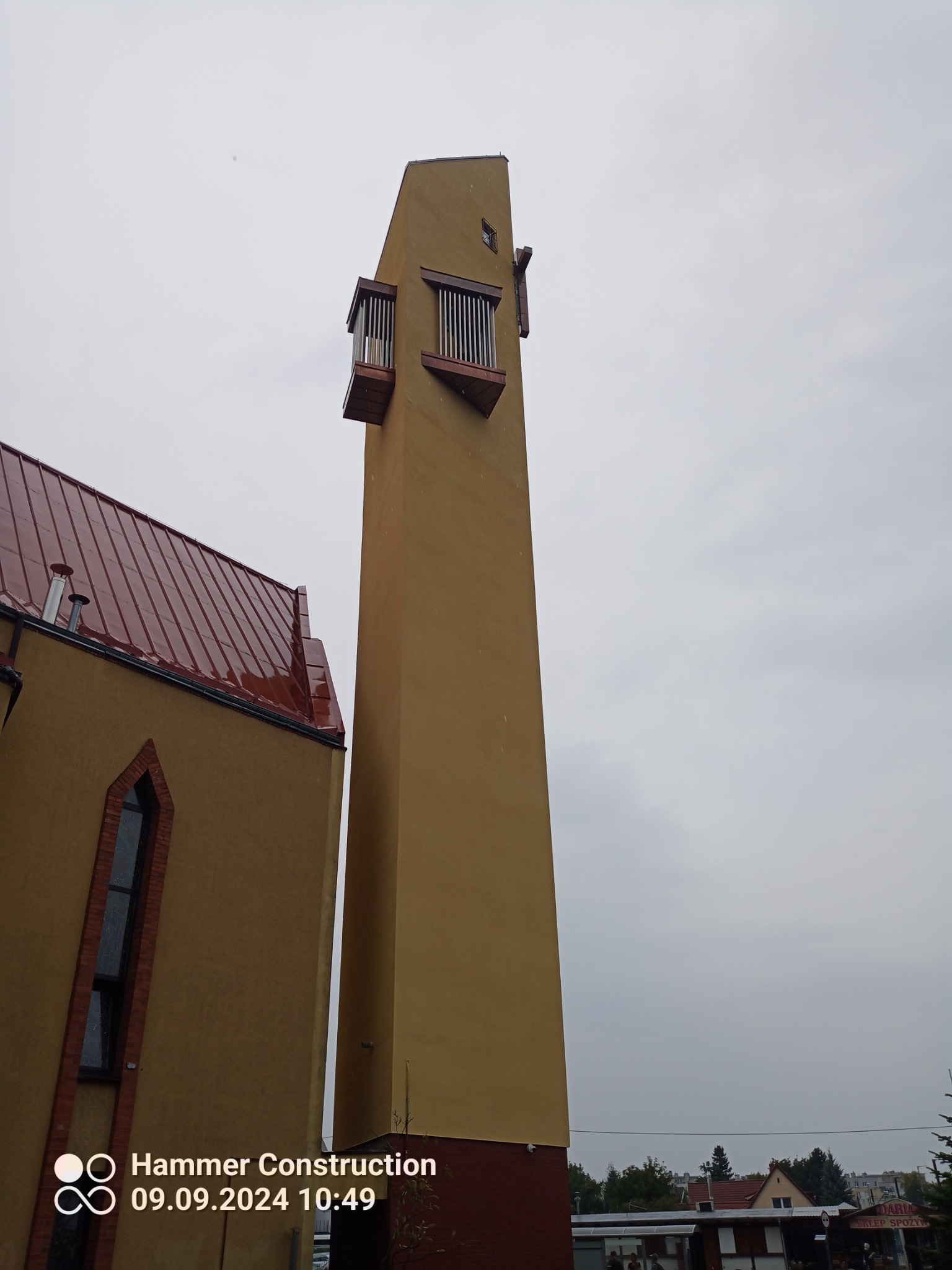 Wieża kościoła w Szczecinie z pomalowaną elewacją w odcieniach beżu i brązu, widoczna z dołu pod pochmurnym niebem.