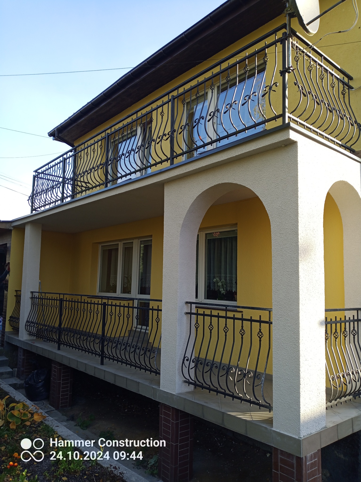 Dwukondygnacyjny dom z żółtą elewacją, białymi kolumnami i czarnymi, ozdobnymi balustradami balkonów. Widoczne dwa balkony, dolny z łukami i górny z satelitą na dachu.